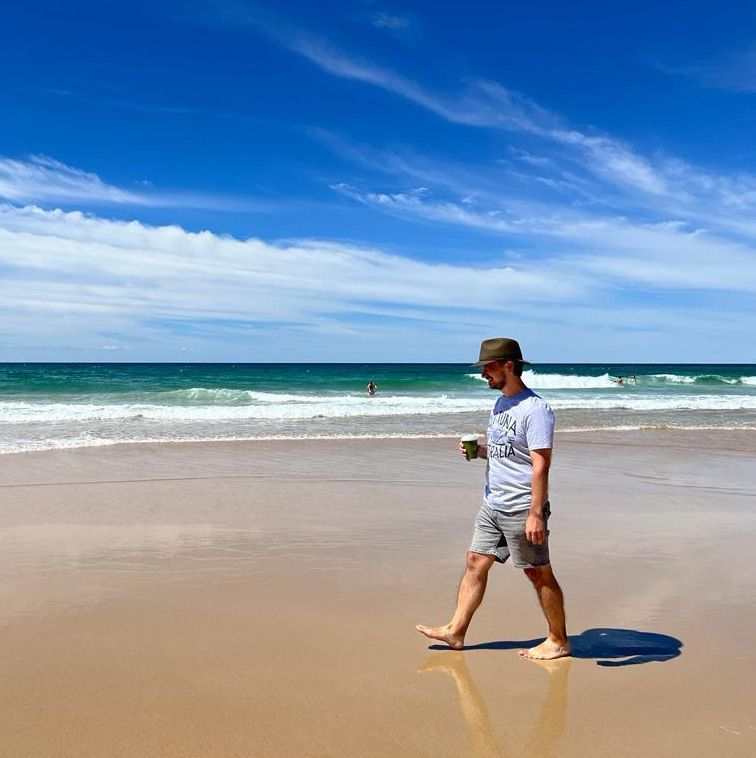 Me walking barefoot along a beach in Australia, wearing a hat and holding a coffee.